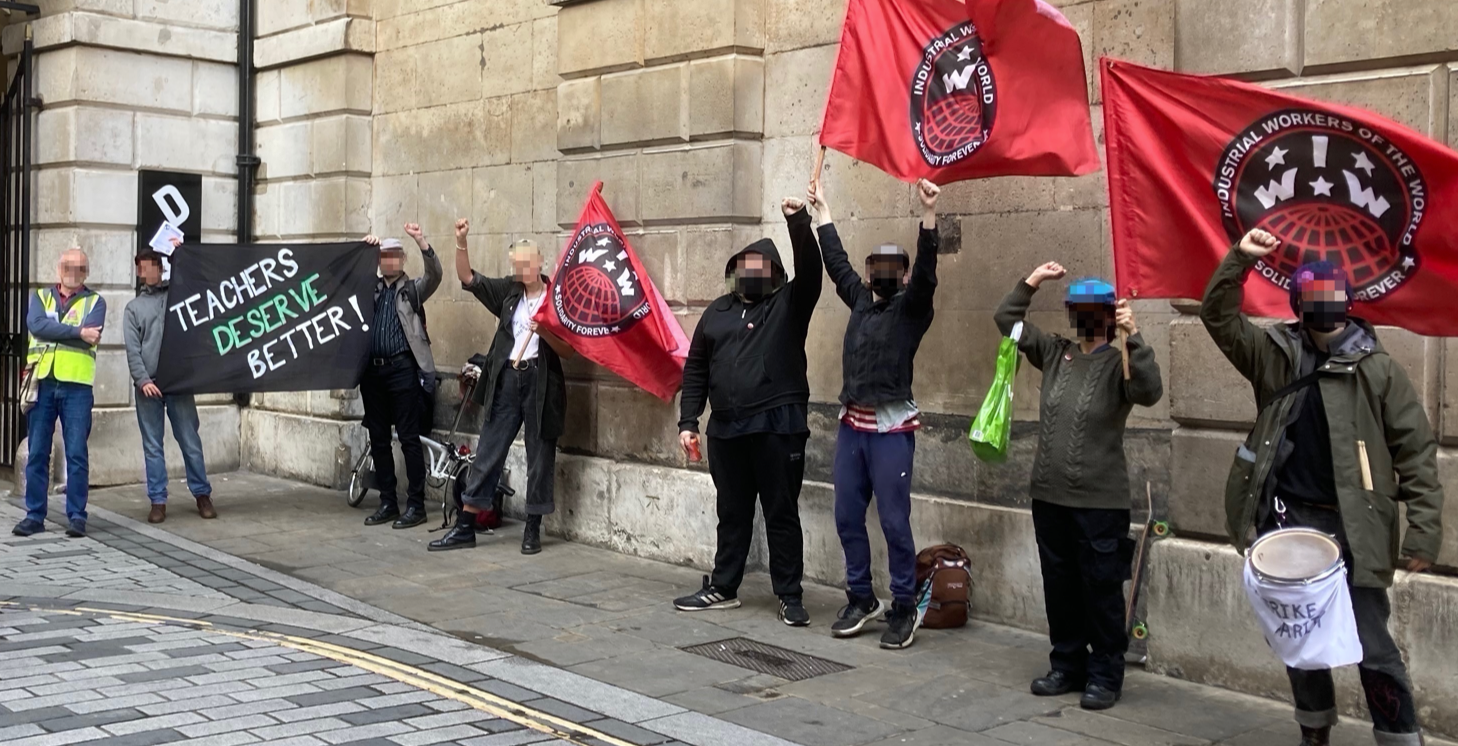Image shows approximately 10 picketers with rep IWW flags standing in front of an arch near Liverpool St Station