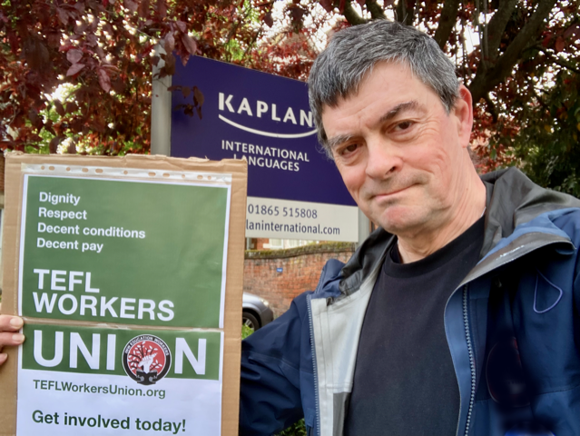 Image shows a man with a placard advertising the TEFL Workers Union. In the background, a sign for Kaplan language school can be seen.