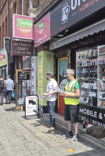 Image shows two men with leaflet, one in a hi viz vest, in front of TopUp English in Camden, London.